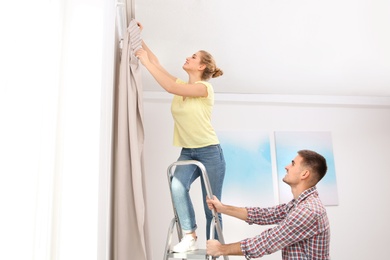 Young couple hanging window curtain indoors. Interior decor element