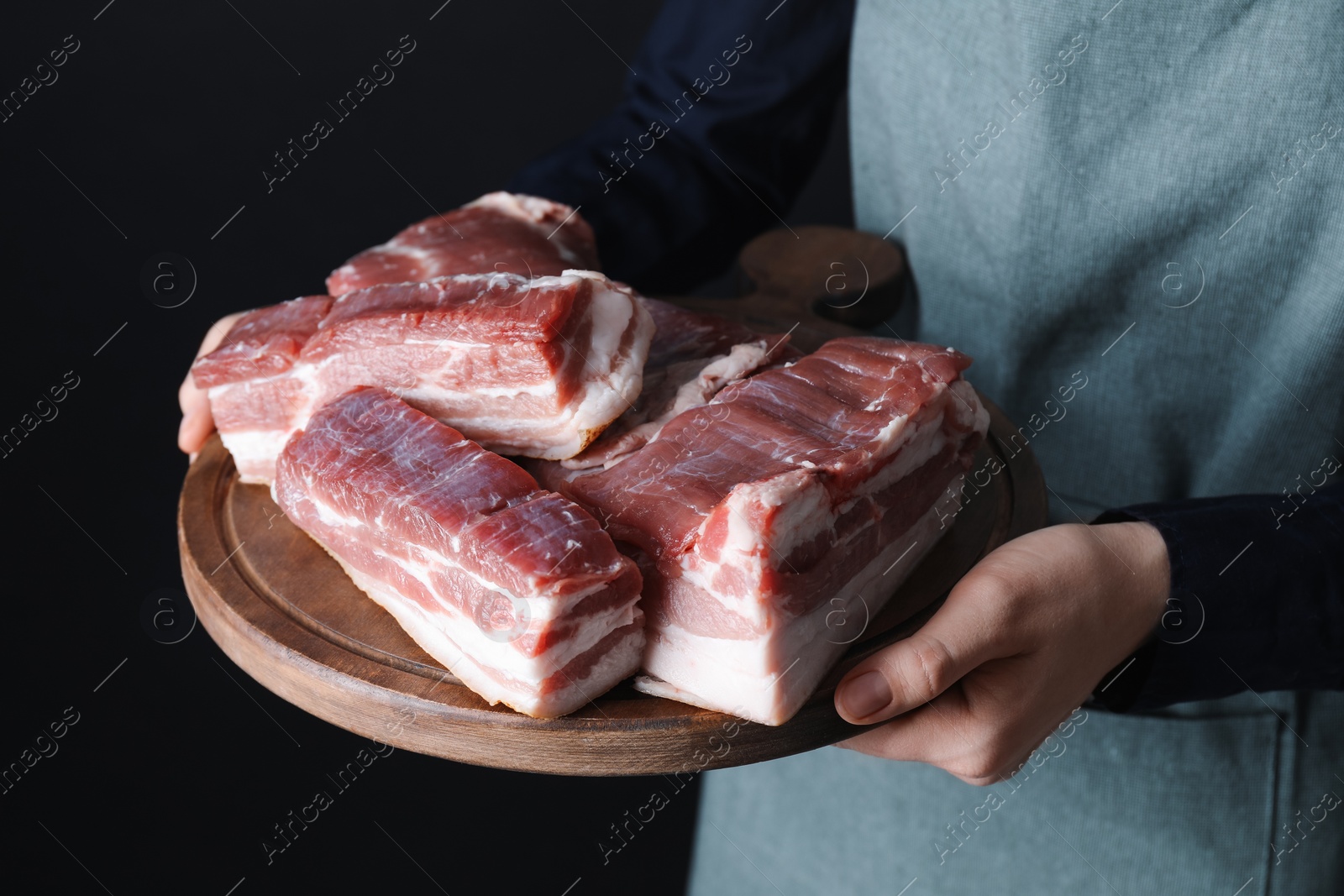 Photo of Woman holding wooden board with pieces of raw pork belly on black background, closeup