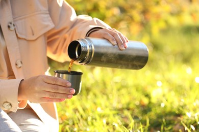 Photo of Woman pouring tea from thermos into cup lid on green grass outdoors, closeup