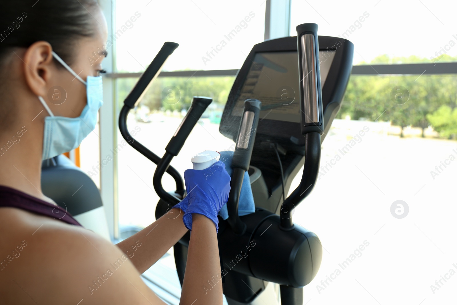 Photo of Woman cleaning exercise equipment with disinfectant spray and cloth in gym