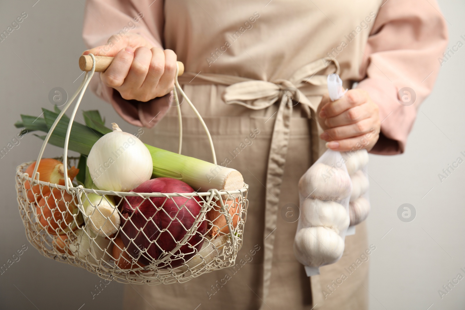 Photo of Woman holding basket with fresh onion bulbs, leeks and garlic on light background, closeup