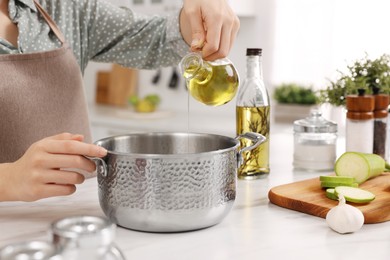 Photo of Cooking process. Woman pouring oil from bottle into pot at white countertop in kitchen, closeup