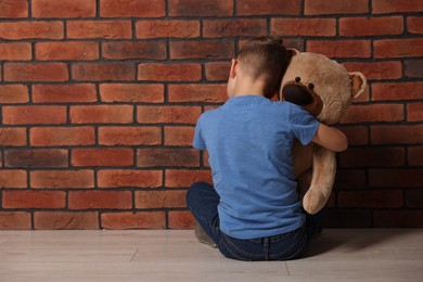 Photo of Child abuse. Upset boy with teddy bear sitting on floor near brick wall indoors, back view. Space for text