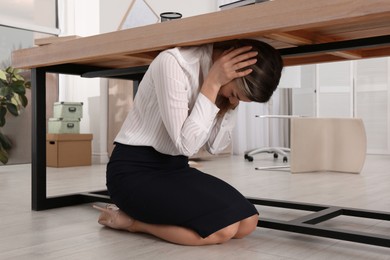 Scared young woman hiding under office desk during earthquake