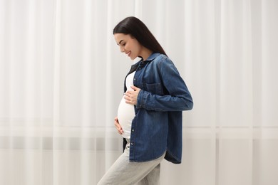 Photo of Happy pregnant woman touching her belly near window indoors