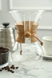 Photo of Empty glass chemex coffeemaker and beans on white marble table, closeup