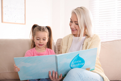 Photo of Mature woman with her little granddaughter reading book together at home