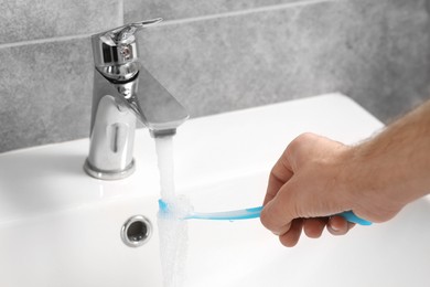 Man holding toothbrush under flowing water above sink in bathroom, closeup