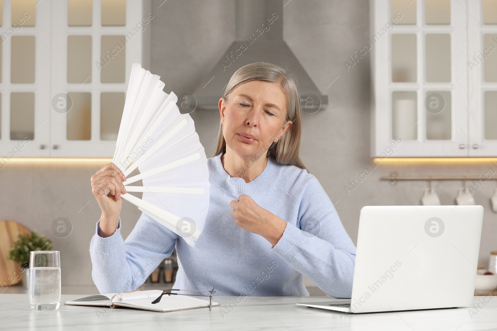 Photo of Menopause. Woman waving hand fan to cool herself during hot flash at table in kitchen