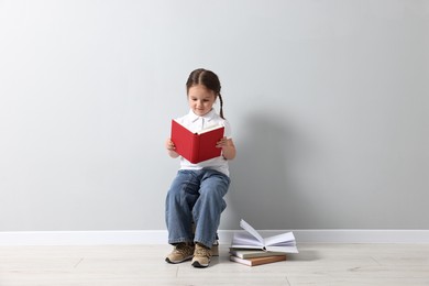 Photo of Cute little girl reading on stack of books near light grey wall