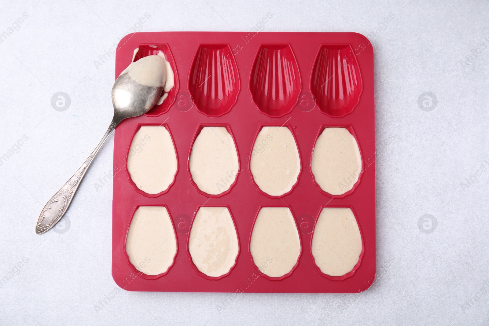 Photo of Baking mold for madeleine cookies with batter and spoon on white table, top view