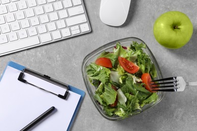 Photo of Fresh vegetable salad, apple, fork, clipboard and keyboard on grey table at workplace, flat lay. Business lunch