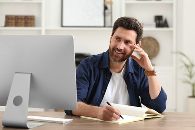 Photo of Home workplace. Happy man taking notes while working with computer at wooden desk in room