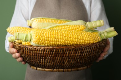 Woman holding wicker basket with tasty fresh corn cobs on green background, closeup