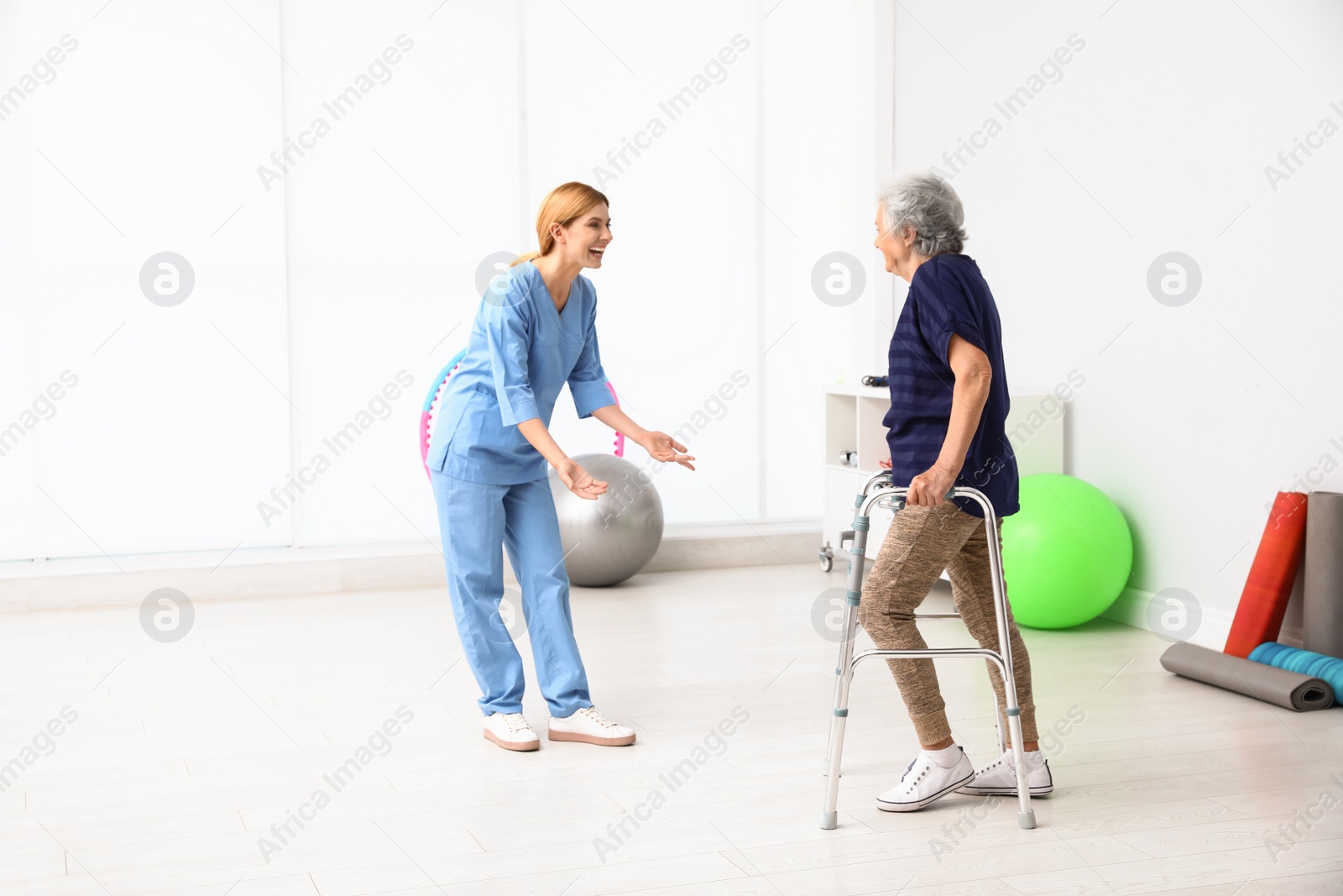 Photo of Caretaker helping elderly woman with walking frame indoors