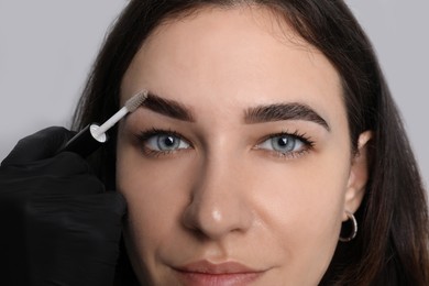 Photo of Beautician fixing woman's eyebrows with gel after tinting on light grey background, closeup
