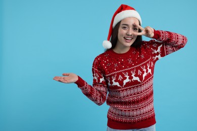 Photo of Happy young woman in Christmas sweater and Santa hat showing something on light blue background. Space for text