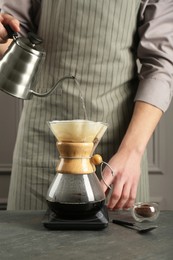 Photo of Man pouring hot water into glass chemex coffeemaker with paper filter and coffee at gray table, closeup