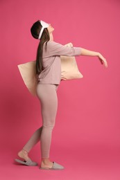 Young woman wearing pajamas, mask and slippers with pillow in sleepwalking state on pink background