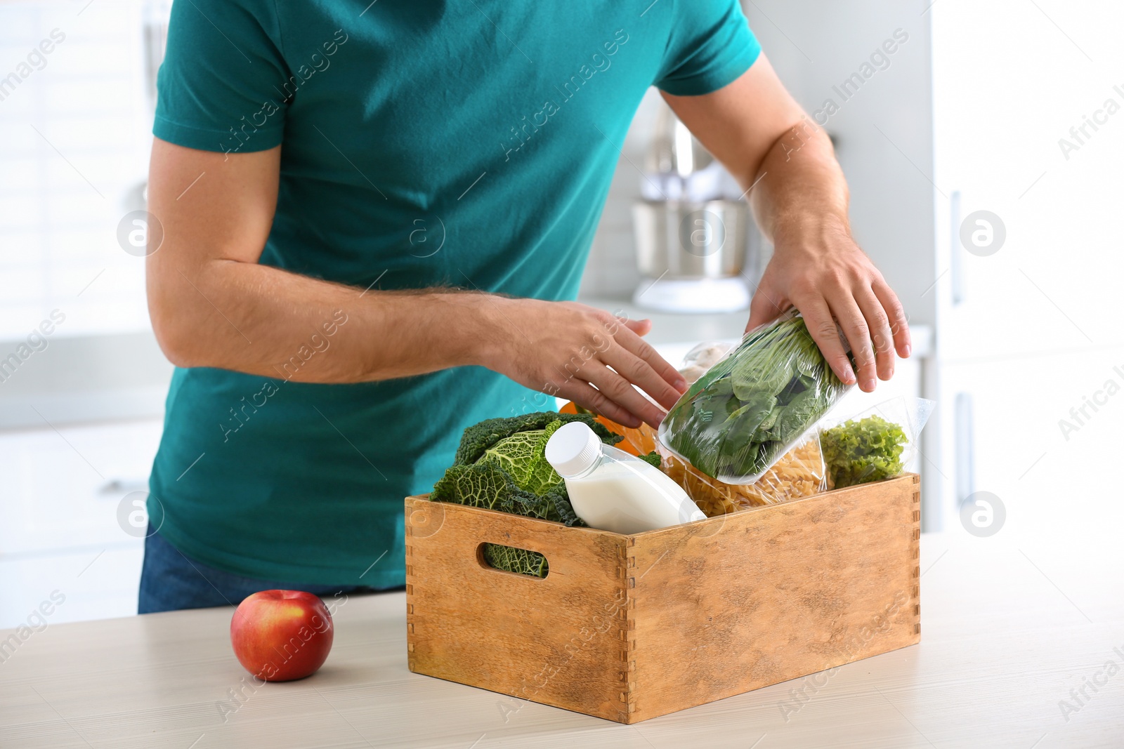 Photo of Man with wooden crate full of products at table in kitchen, closeup. Food delivery service