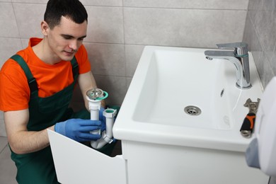 Young plumber wearing protective gloves repairing sink in bathroom