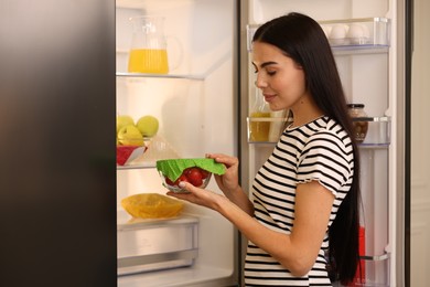 Photo of Woman holding bowl of fresh tomatoes covered with beeswax food wrap near refrigerator indoors