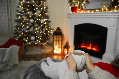Photo of Woman with hot drink resting near fireplace in cozy room decorated for Christmas, closeup