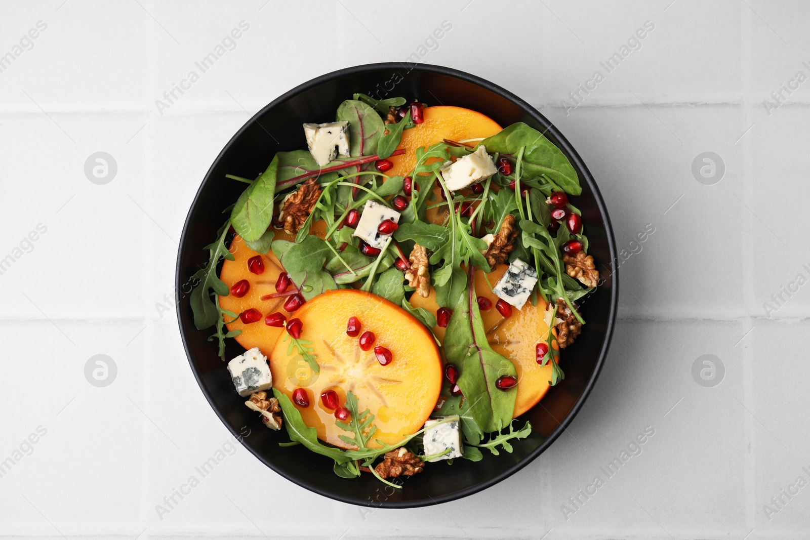 Photo of Tasty salad with persimmon, blue cheese, pomegranate and walnuts served on white tiled table, top view