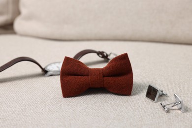 Stylish brown bow tie, wristwatch and cufflinks on light grey sofa