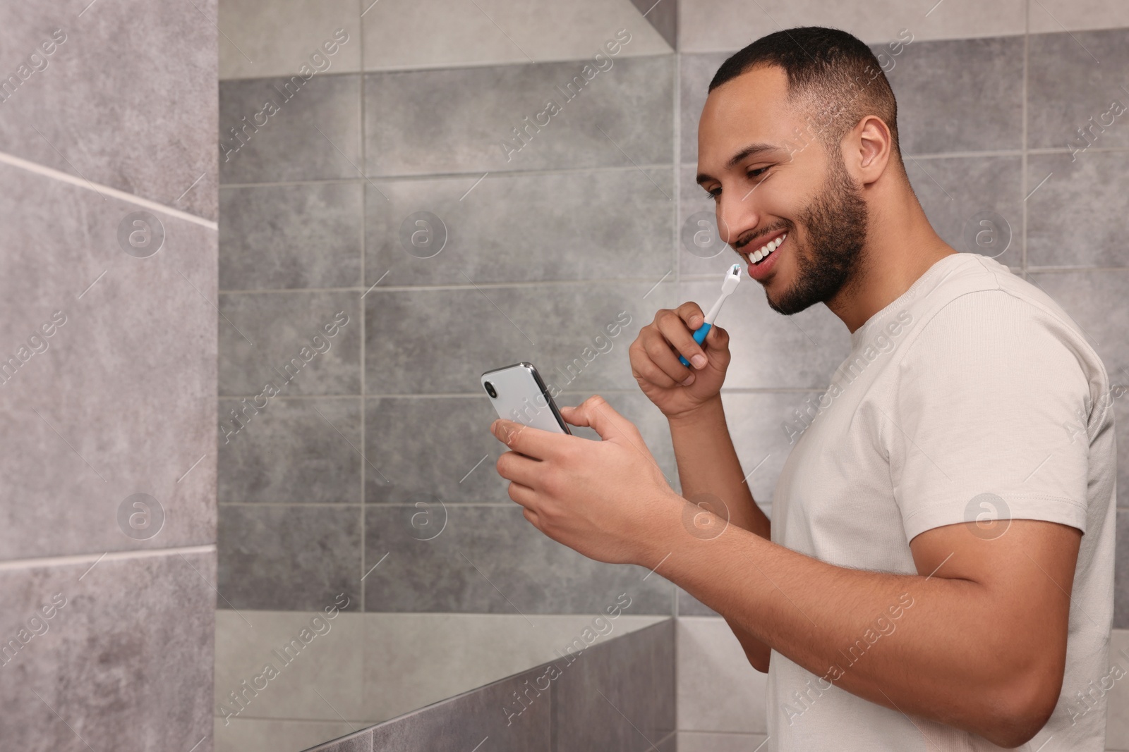 Photo of Happy man using smartphone while brushing teeth in bathroom, space for text. Internet addiction