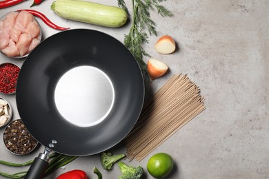 Empty iron wok surrounded by raw ingredients on grey table, flat lay. Space for text