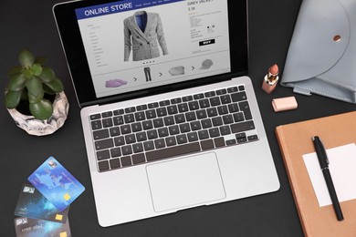 Photo of Online store website on laptop screen. Computer, credit cards, women's bag, stationery and lipstick on black background, above view