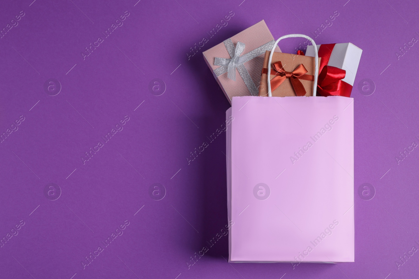 Photo of Pink paper shopping bag full of gift boxes on purple background, flat lay. Space for text