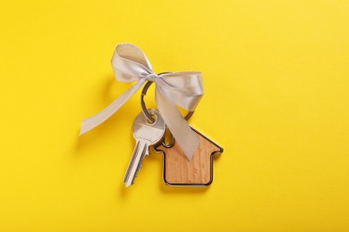 Key with trinket in shape of house and grey bow on yellow background, top view. Housewarming party