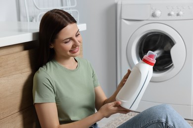 Photo of Woman sitting near washing machine and holding fabric softener in bathroom