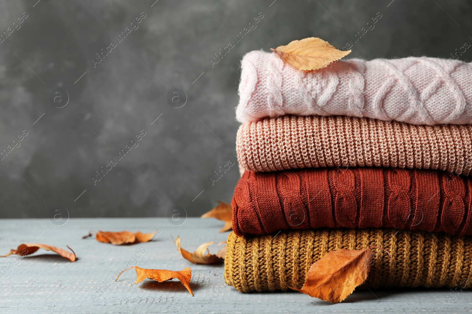 Photo of Stack of warm clothes and autumn leaves on wooden table against grey background. Space for text