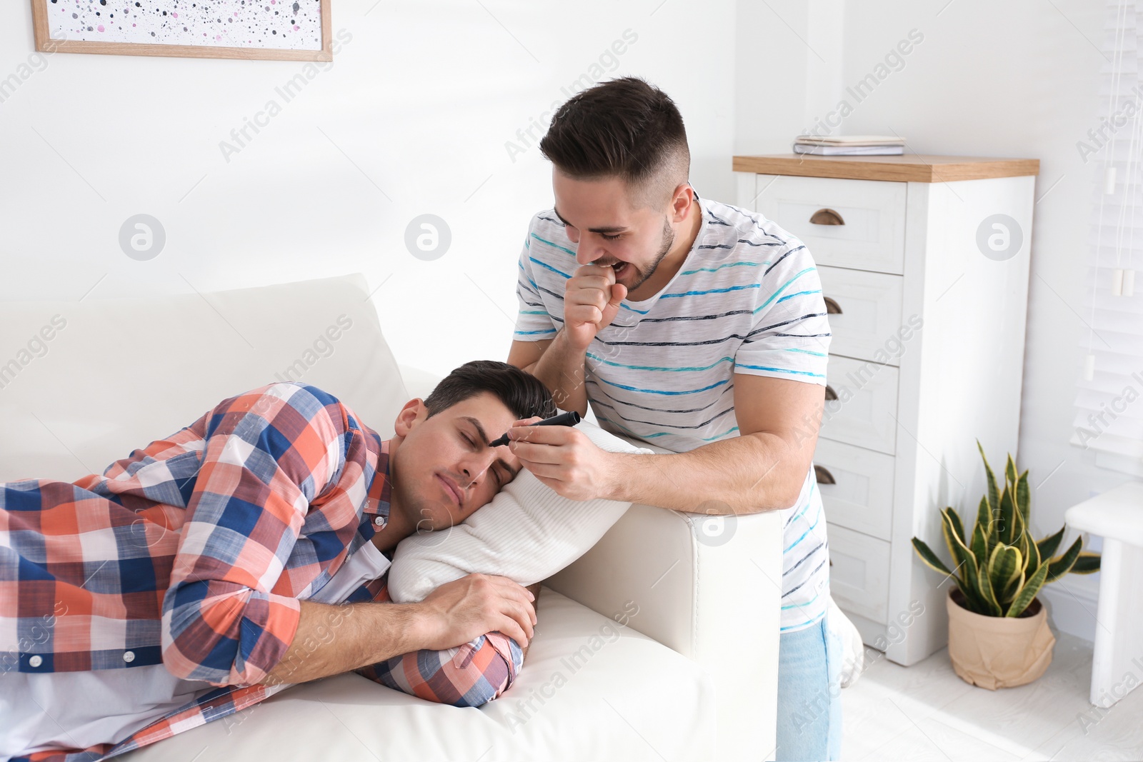 Photo of Young man with marker near sleeping friend indoors. April fool's day