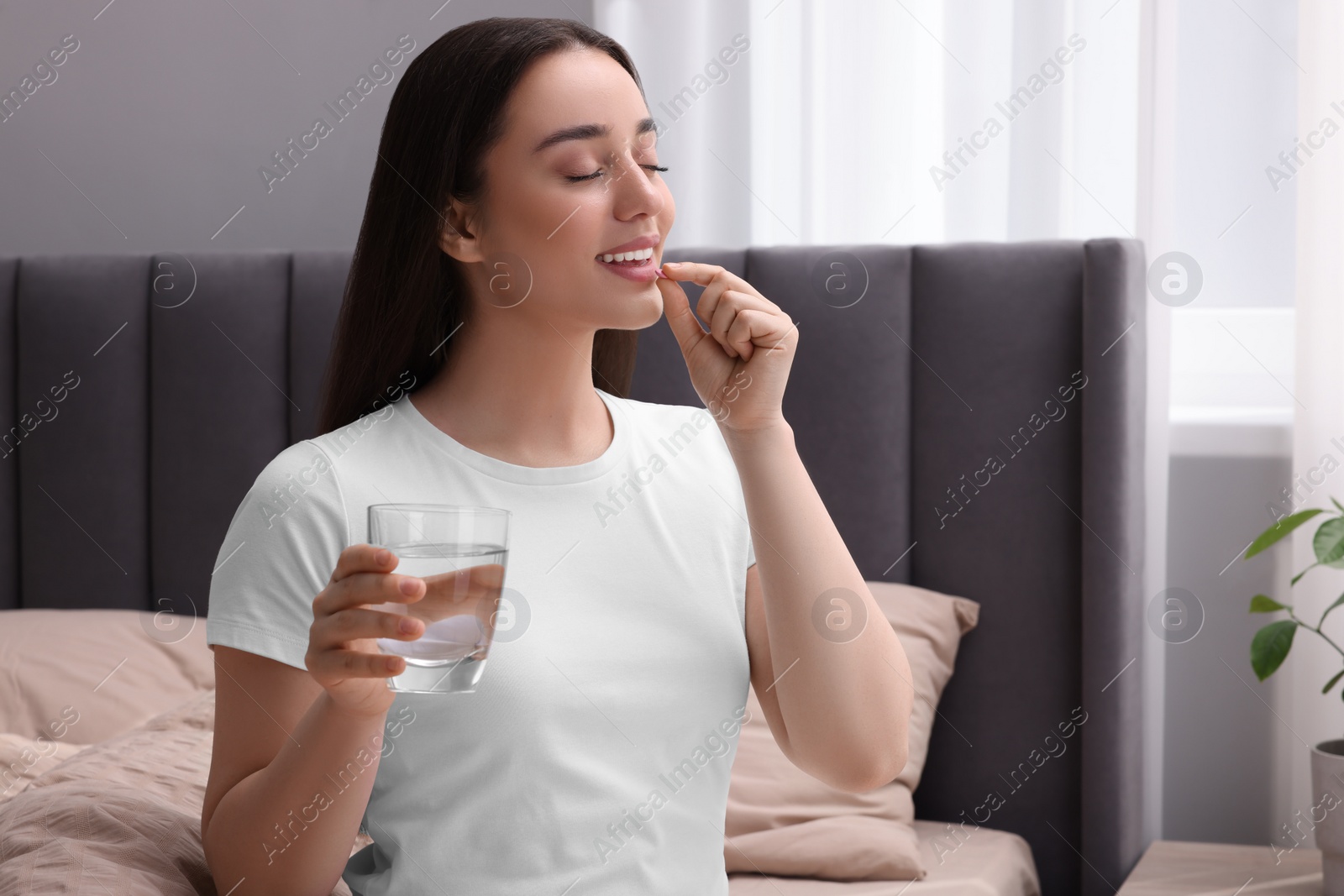 Photo of Beautiful young woman with glass of water taking pill at home, space for text