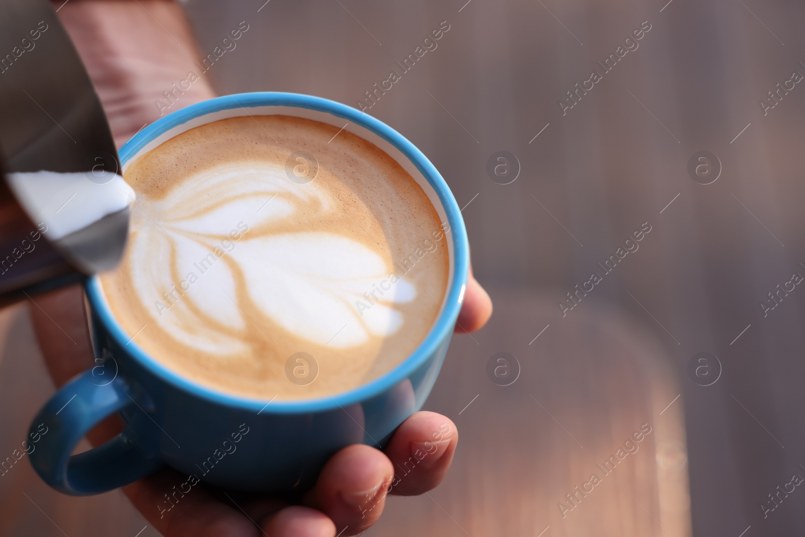 Photo of Barista pouring milk into cup of coffee on blurred background, closeup. Space for text