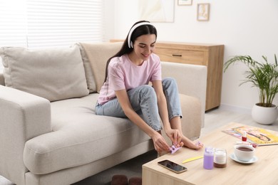 Photo of Beautiful young woman listening to music while preparing toenails for pedicure in living room