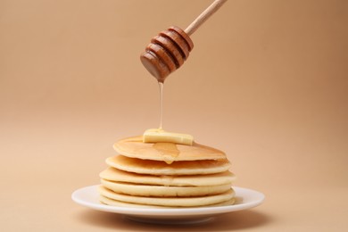 Pouring honey from dipper onto delicious pancakes with butter on beige background