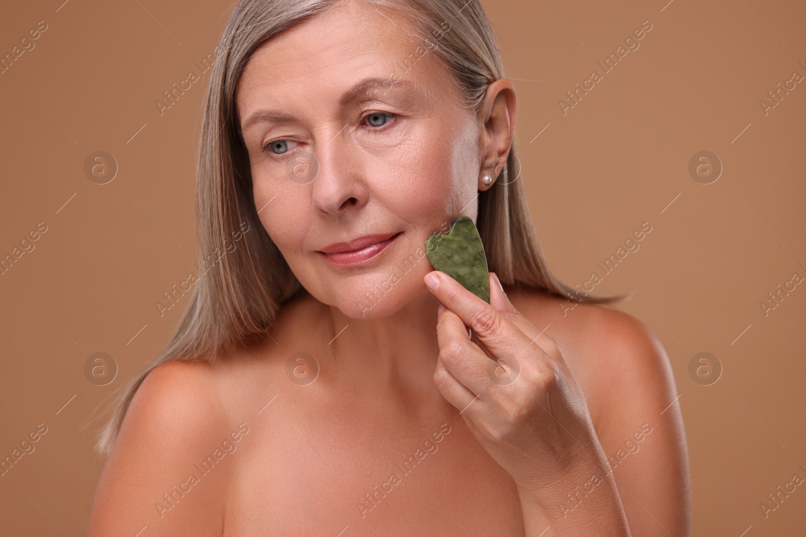 Photo of Woman massaging her face with jade gua sha tool on brown background