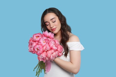 Beautiful young woman with bouquet of pink peonies on light blue background