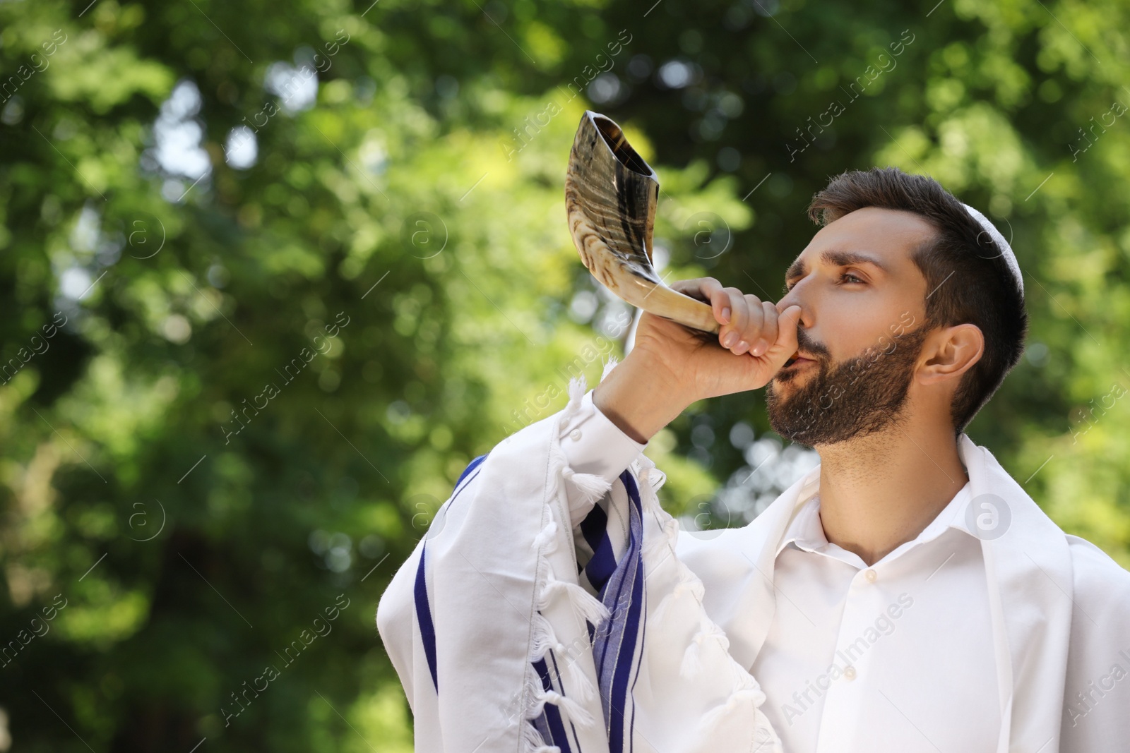 Photo of Jewish man in kippah and tallit blowing shofar outdoors. Rosh Hashanah celebration