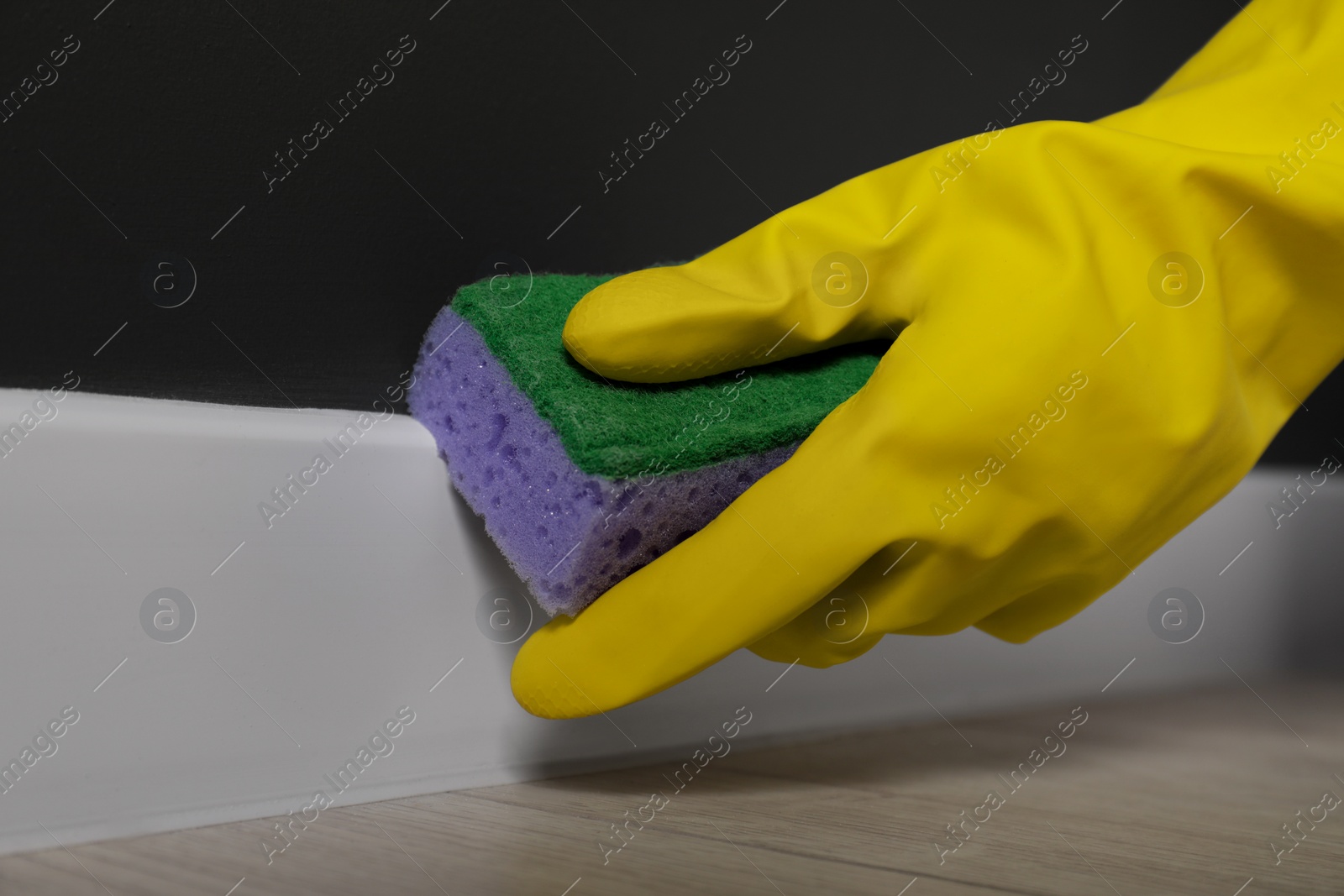 Photo of Woman in protective glove cleaning plinth with sponge indoors, closeup