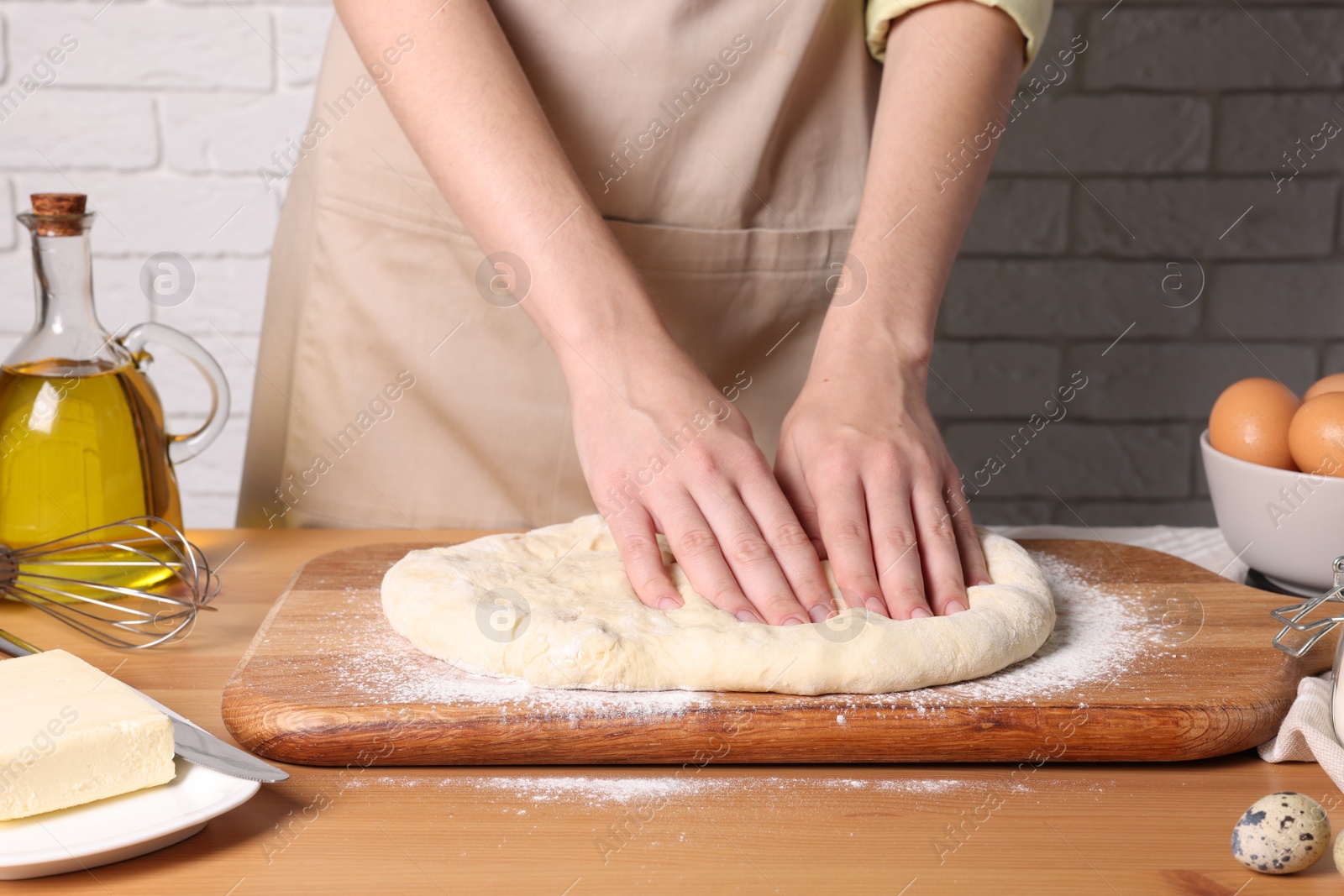 Photo of Woman kneading dough at wooden table near white brick wall, closeup