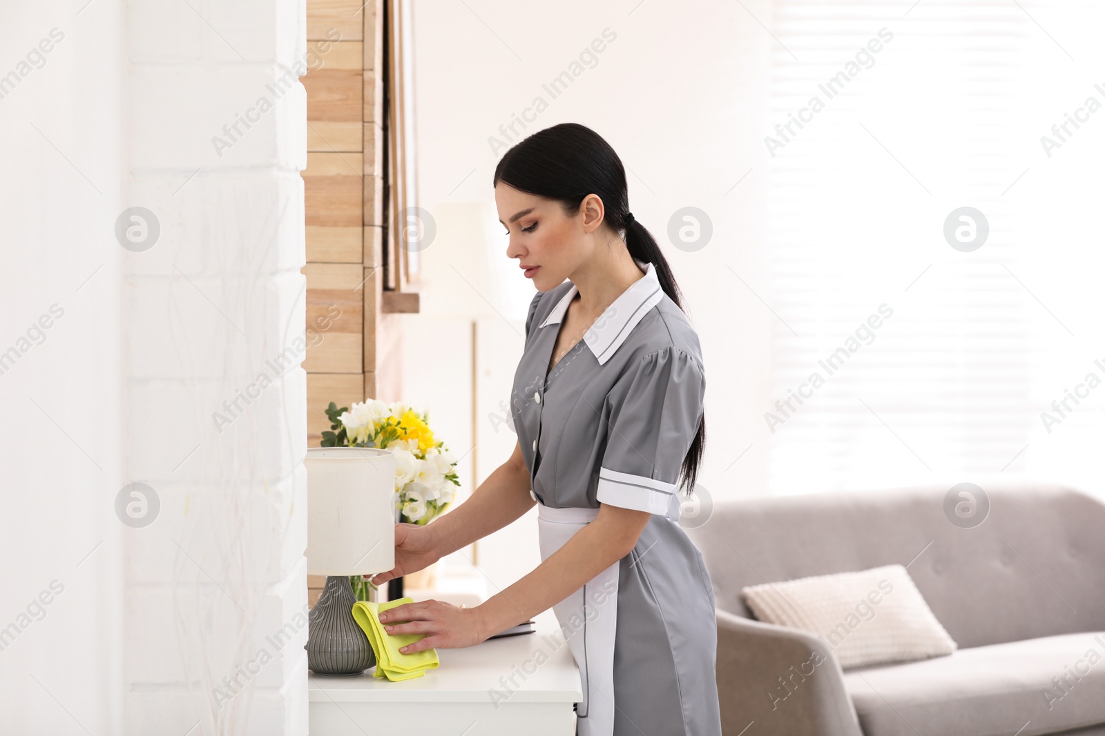 Photo of Young chambermaid wiping dust from furniture in hotel room