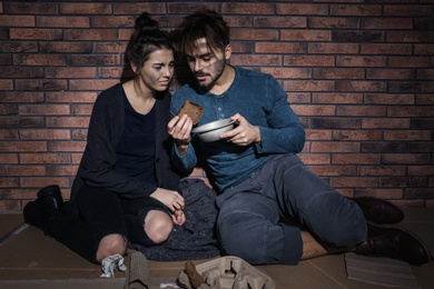 Photo of Poor young couple with bread on floor near brick wall
