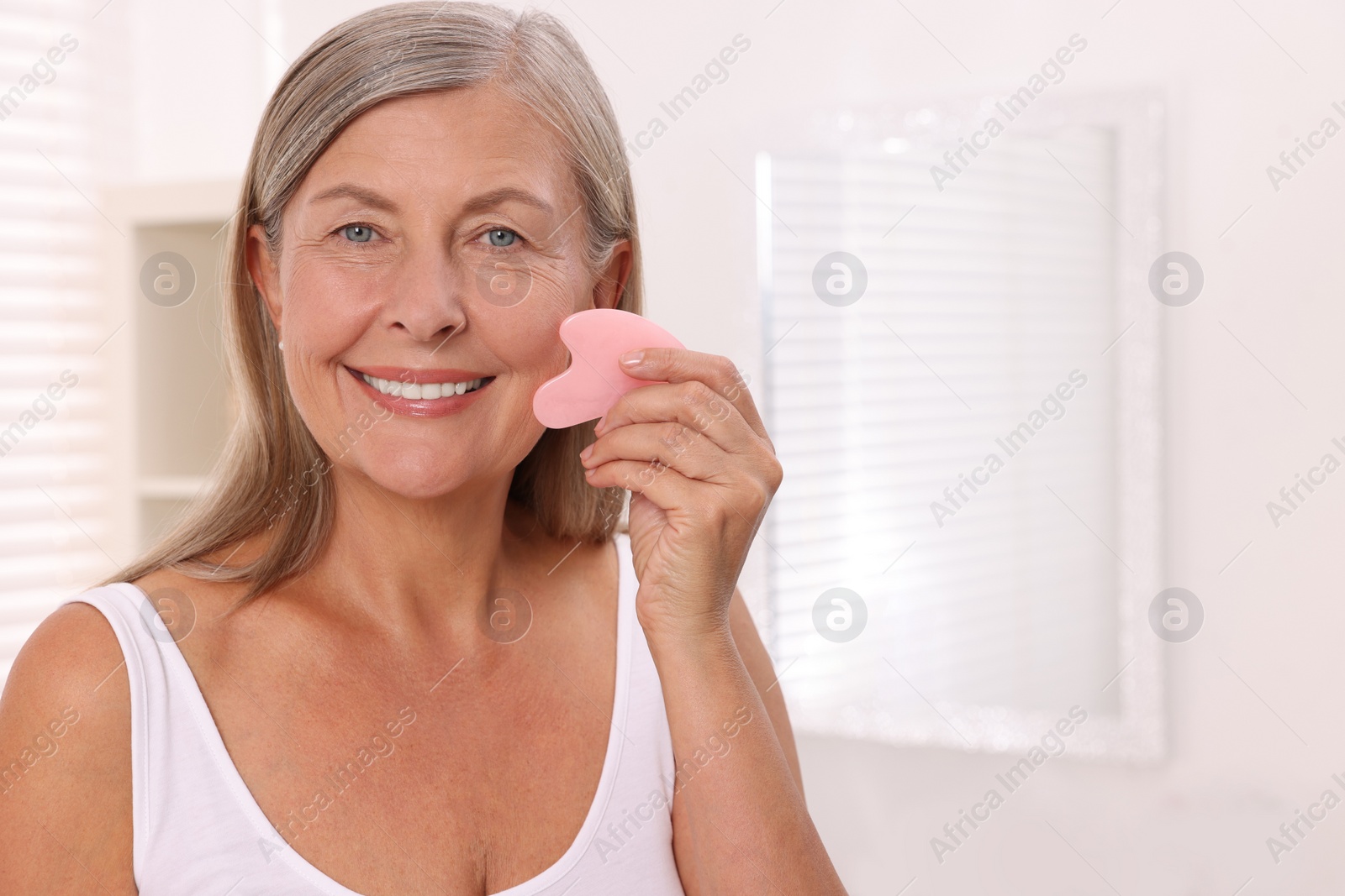 Photo of Woman massaging her face with rose quartz gua sha tool in bathroom. Space for text