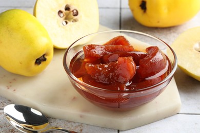 Tasty homemade quince jam in bowl, spoon and fruits on tiled table, closeup